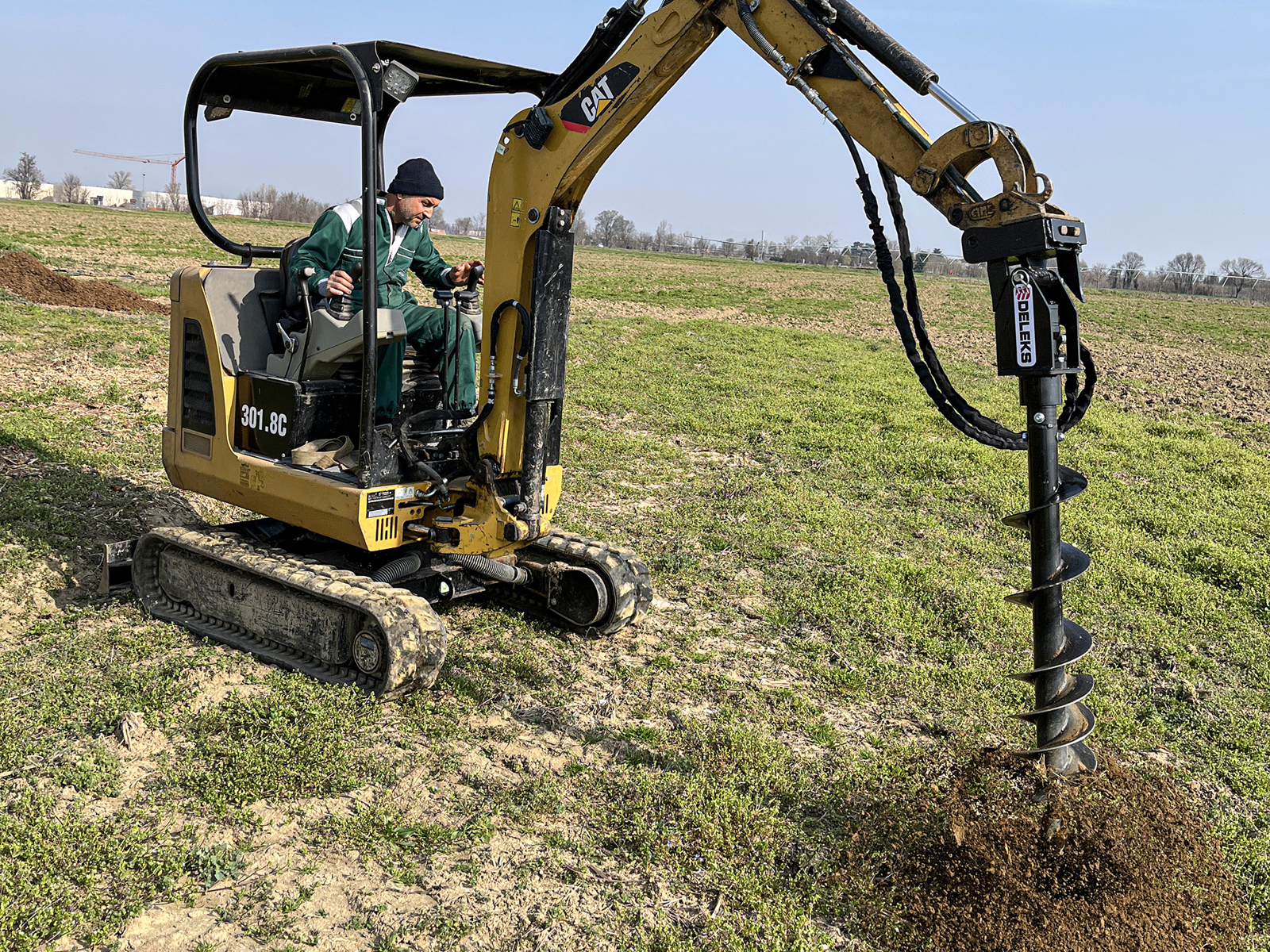 Der Bagger mit Bediener und Erdbohrgerät bohrt ein Loch in den Boden auf einer Wiese